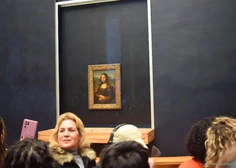 Visitors in front of the Mona Lisa behind glass on its dark gallery wall, a highlight with Louvre Museum tickets