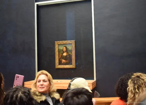 Zwiedzający przed Mona Lisą za szybą na ciemnej ścianie sali, punkt kulminacyjny wycieczki z biletami do Luwru